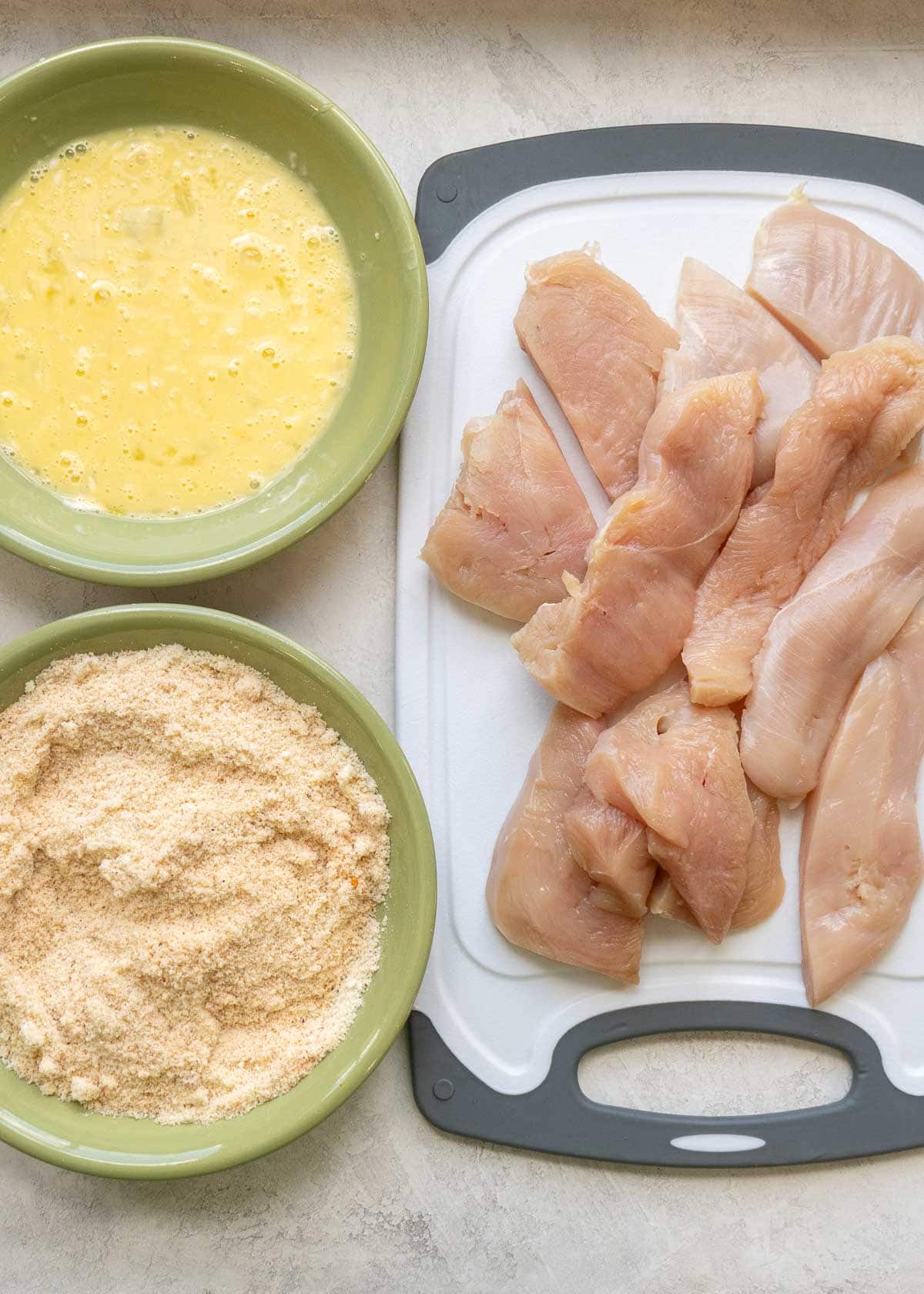 egg and mayonnaise mixture in bowl #1, almond flour, parmesan, and seasoning mixture in bowl #2, and sliced chicken tenders on a cutting board