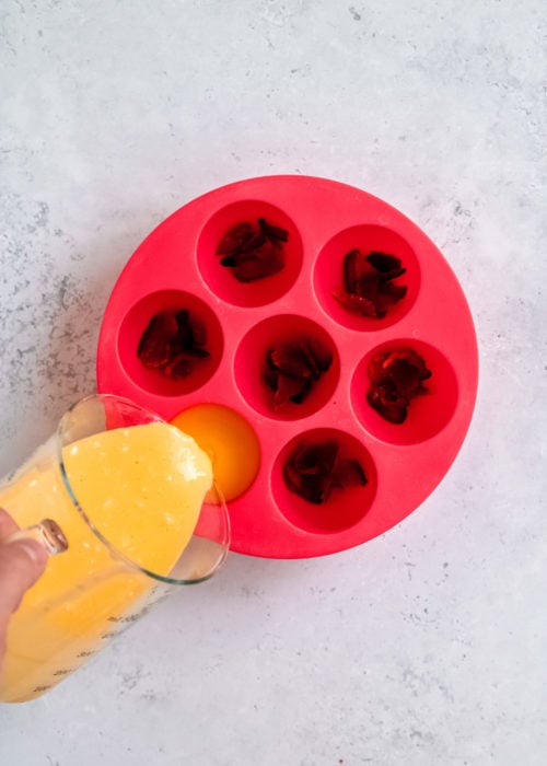 crumbled bacon in each cavity of a red 7-egg-bite mold. The whisked egg mixture is being poured into one cavity.