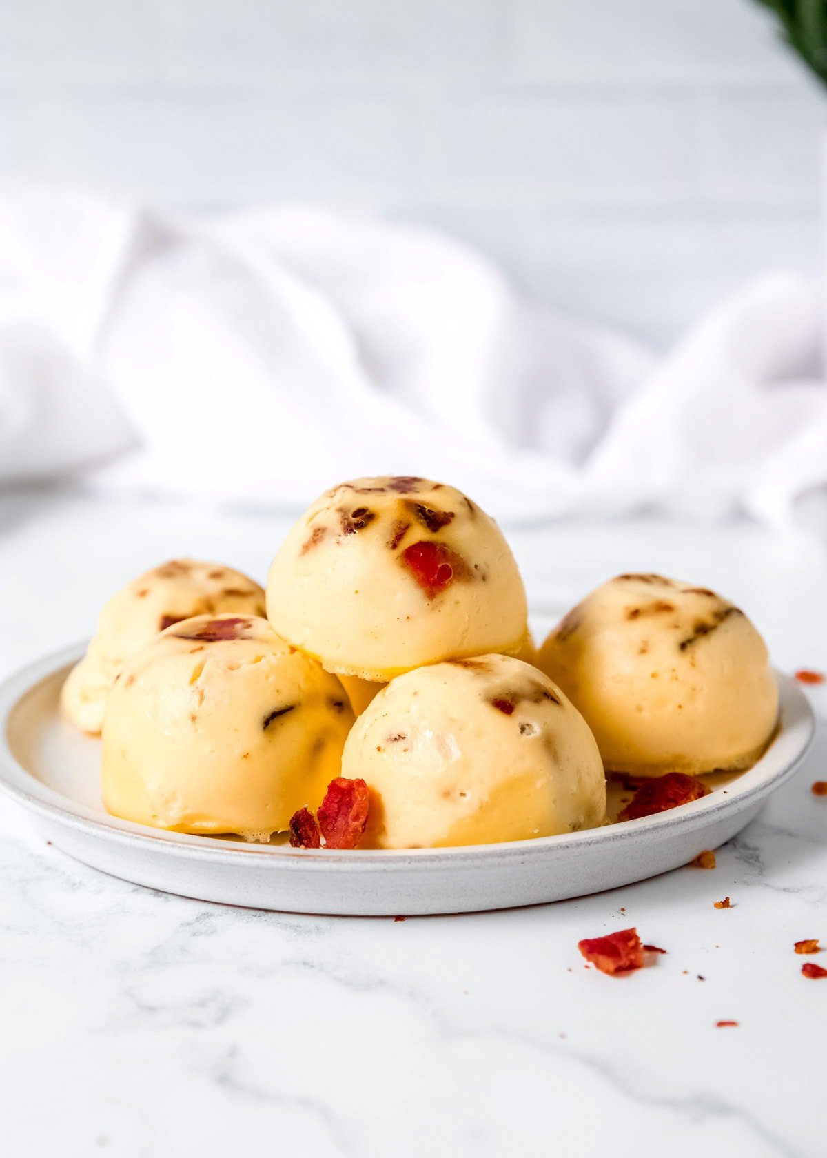 several egg bites piled on a white plate with bacon scattered on the counter