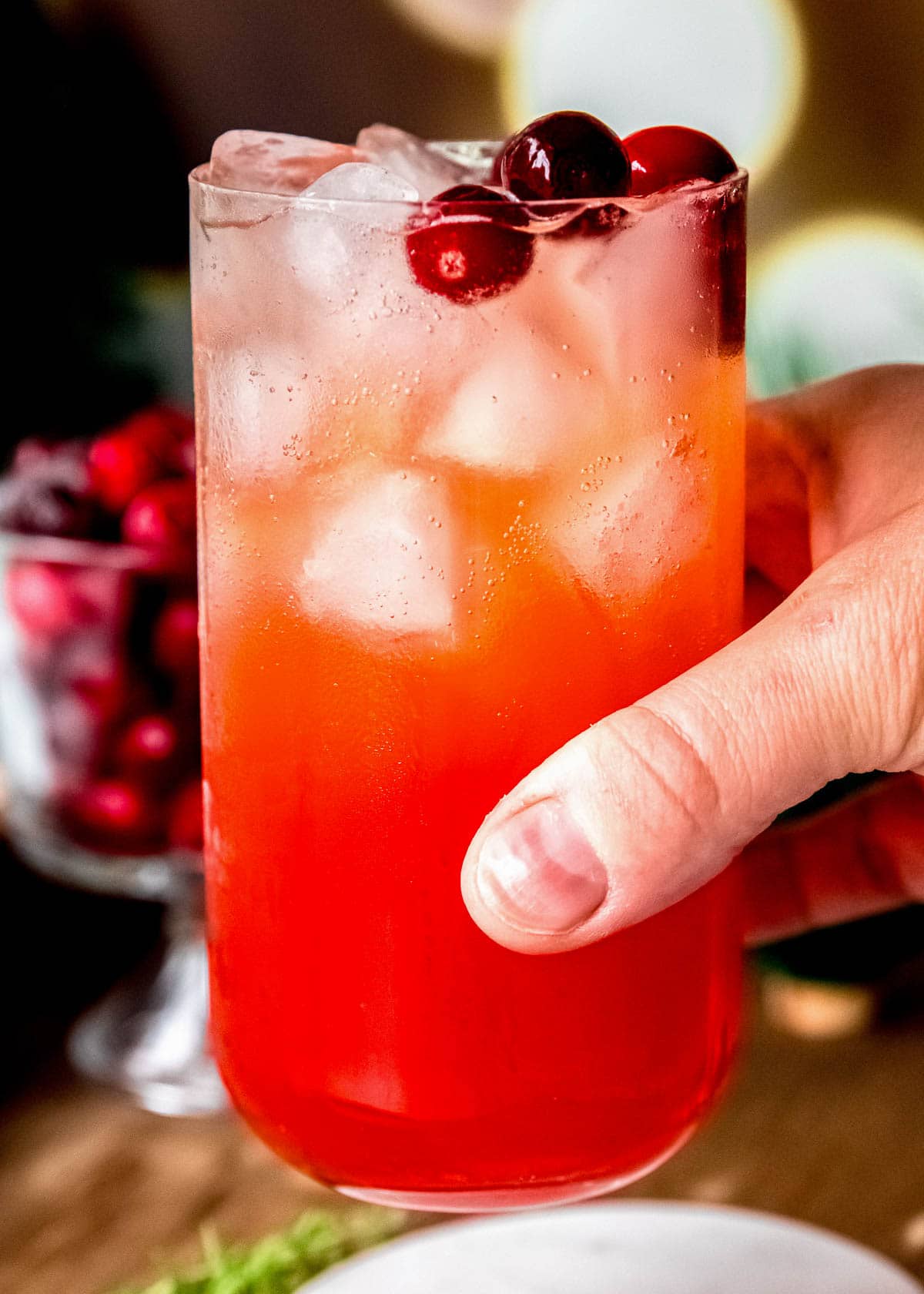 hand grabbing a cranberry aperol spritz with cranberries and thyme in the background
