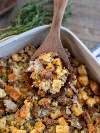an overhead shot of sausage stuffing in a dish with a brown spoon and herbs on the side.