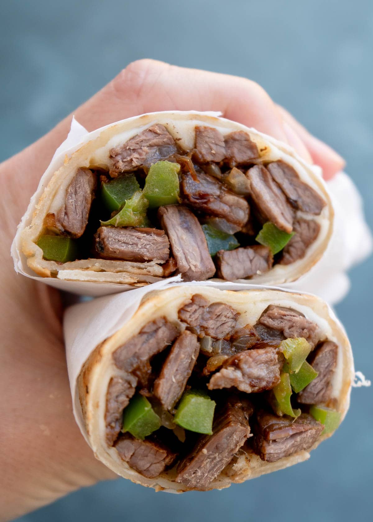 a hand holding two meaty philly cheesesteak wraps full of sirloin, peppers, and onions