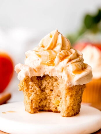an apple cinnamon cupcake topped with caramel buttercream and caramel sauce on a white plate; a bite has been taken from the cupcake showing the fluffy texture of the spiced fall cupcake