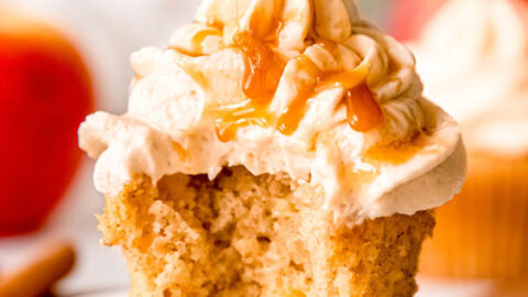 a gluten-free apple cinnamon cupcake on a white plate topped with a tall swirl of caramel buttercream being drizzled with extra caramel sauce; a bite has been bitten out of the cupcake to show the fluffy inside.