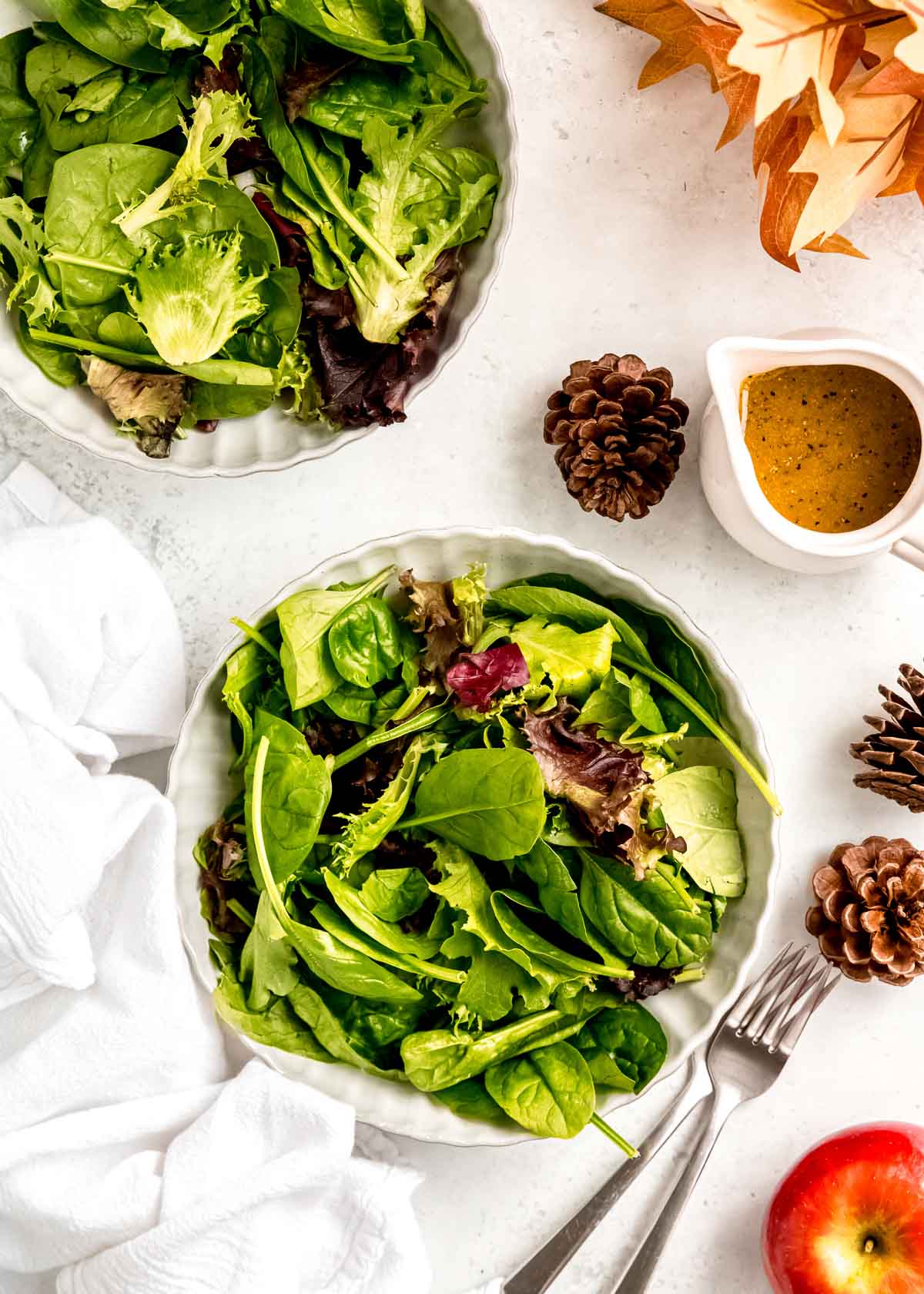 two bowls of mixed greens on a table beside a pitcher of maple-dijon dressing