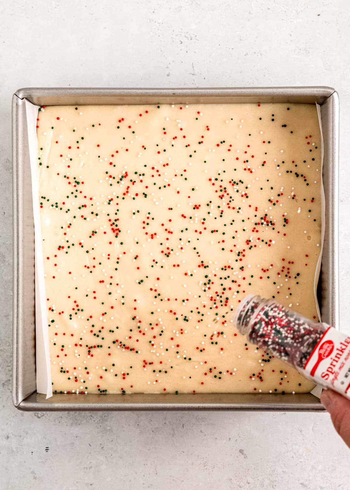 christmas sprinkles being poured over sugar cookie fudge in a pan lined with parchment paper