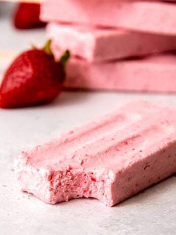 A delicious strawberry popsicle with a bite taken out; in the background there is a stack of more popsicles