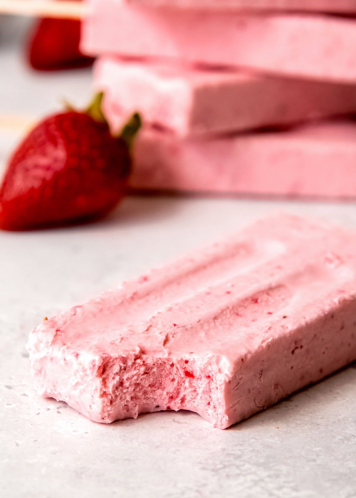 A delicious strawberry popsicle with a bite taken out; in the background there is a stack of more popsicles
