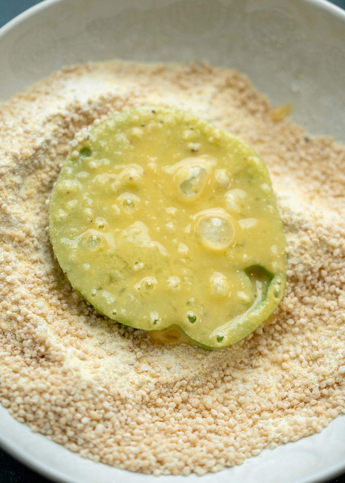 green tomato slice covered in egg wash, laying in a bowl of cornmeal breading