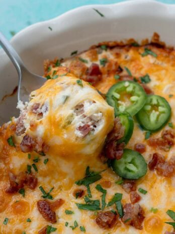 a spoon scooping out some jalapeno popper dip topped with cheese, jalapenos, chives, and bacon