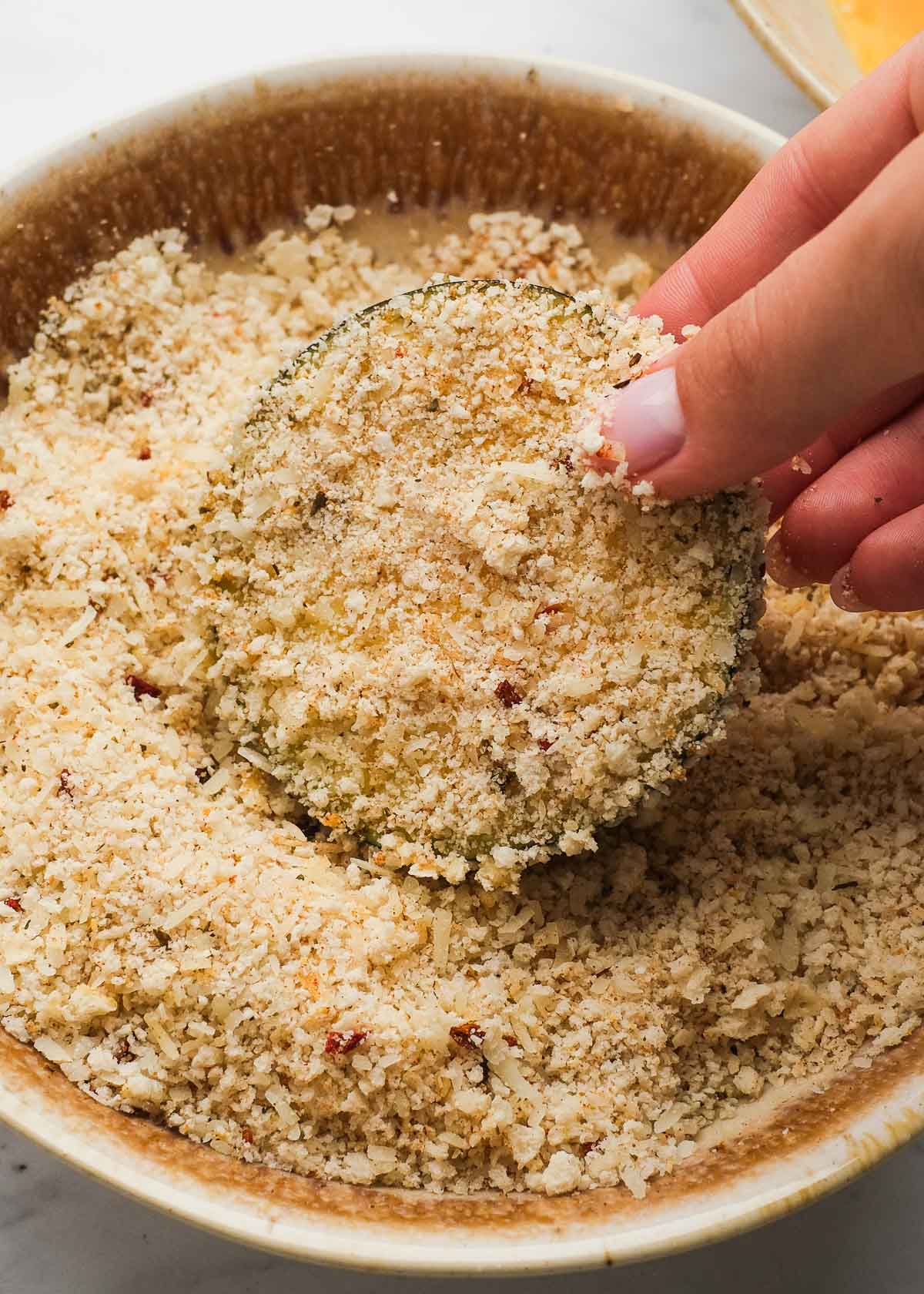 A slice of zucchini being dipped in a seasoned parmesan breadcrumb mixture after being dipped in egg wash.