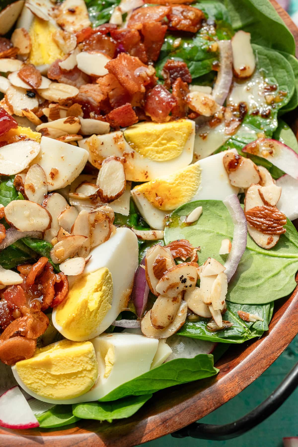 closeup of spinach salad dressing over salad with hard boiled eggs, bacon, radishes, almonds, and more
