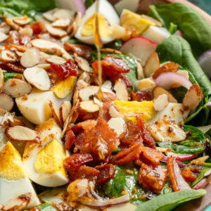 a bowl full of spinach salad topped with hard boiled eggs, crunchy almonds, crispy bacon, and a drizzle of dressing