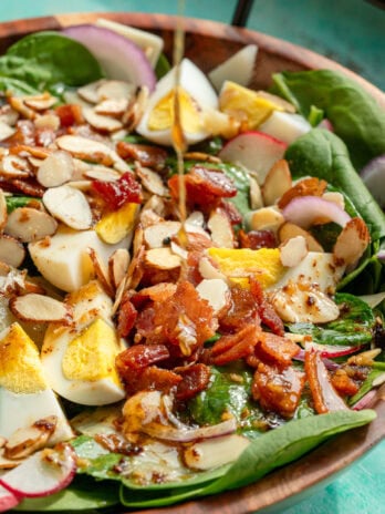 a bowl full of spinach salad topped with hard boiled eggs, crunchy almonds, crispy bacon, and a drizzle of dressing