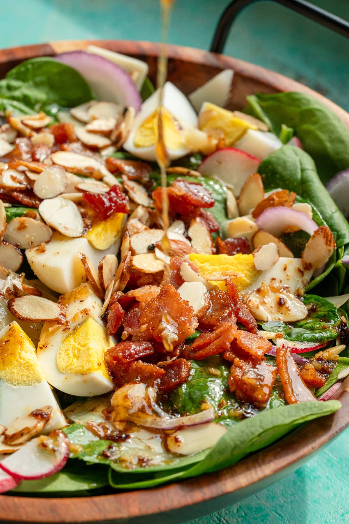 a bowl full of spinach salad topped with hard boiled eggs, crunchy almonds, crispy bacon, and a drizzle of dressing