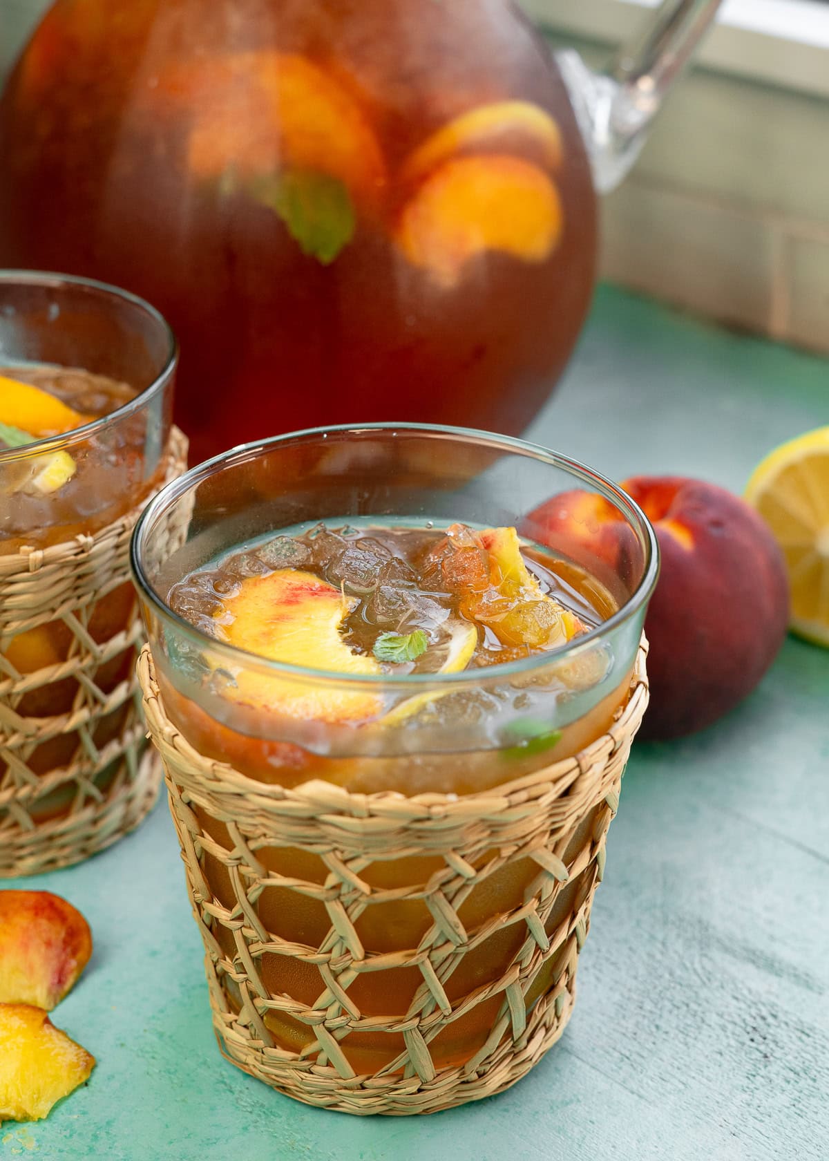 two glasses of peach tea made with peach syrup; behind is a large pitcher garnished with peach slices and mint