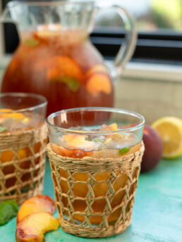 two glasses of peach tea on a table with peaches and a large pitcher