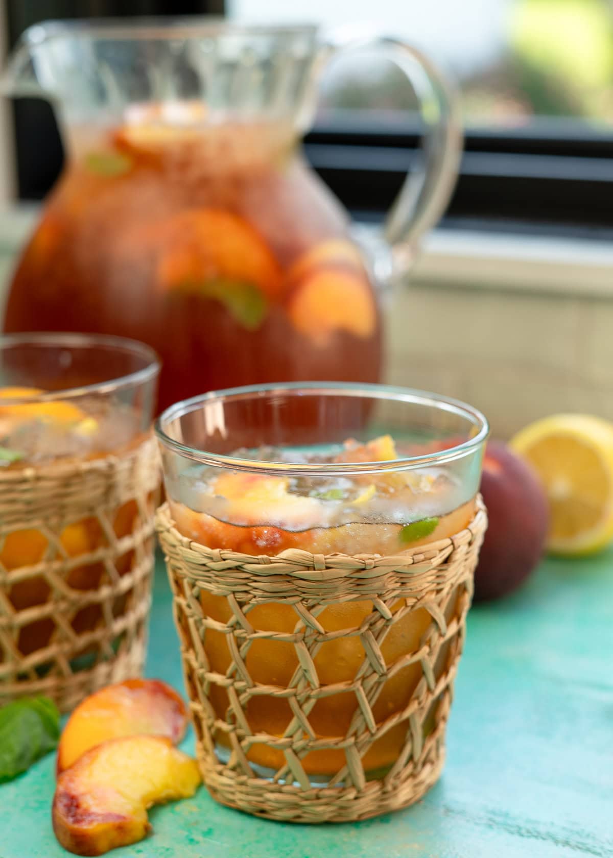 two glasses of peach tea on a table with peaches and a large pitcher