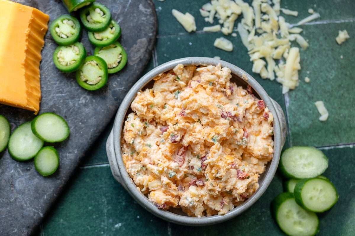 a bowl full of the best pimento cheese dip surrounded by shredded cheese, sliced jalapenos, and sliced cucumber